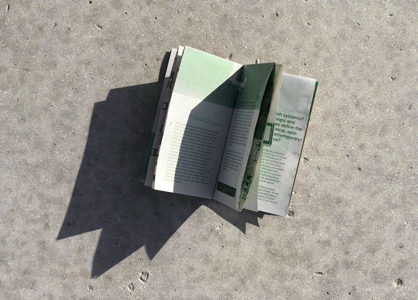 Overhead shot of an open book on a concrete ground. The text and images are green and the inner folds are densely textured with an unidentifiable pattern.